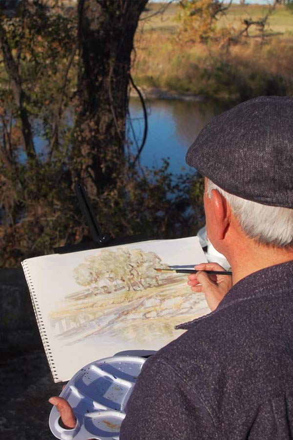 a man doing a watercolor painting outside near a body of water and trees