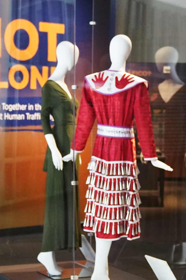 a museum display case containing two mannequins, one wearing a green Dressember dress and the other wearing a Red Jingle Dress by Nan Blassingame