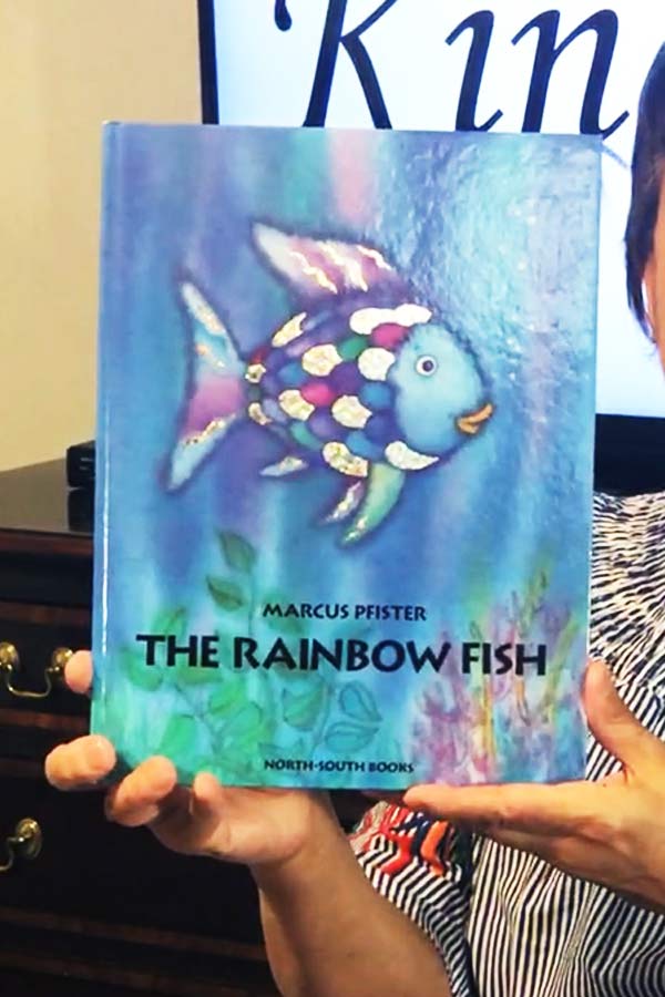 closeup of Museum Educator holding the book titled, "The Rainbow FIsh" by Marcus Pfister which features an illustration of a colorful fish on a blue background