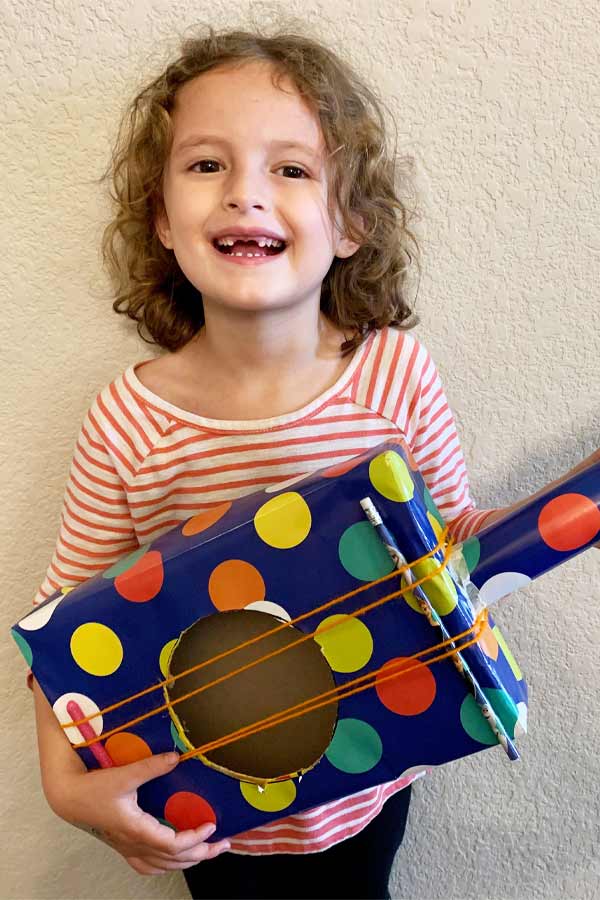 child smiling and holding a "guitar" made out of cardboard and decorated with polka dots 