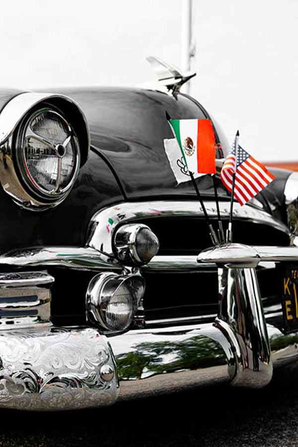 close up of the front of a black lowrider car with silver rims and the Mexican and American flags secured to the front of the car