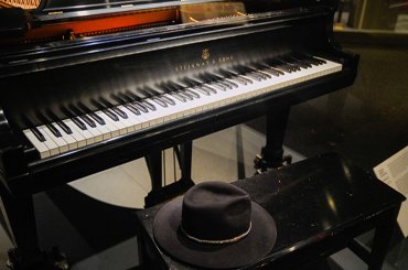 A large black piano, bench, and black fedora hat in a Museum display case.