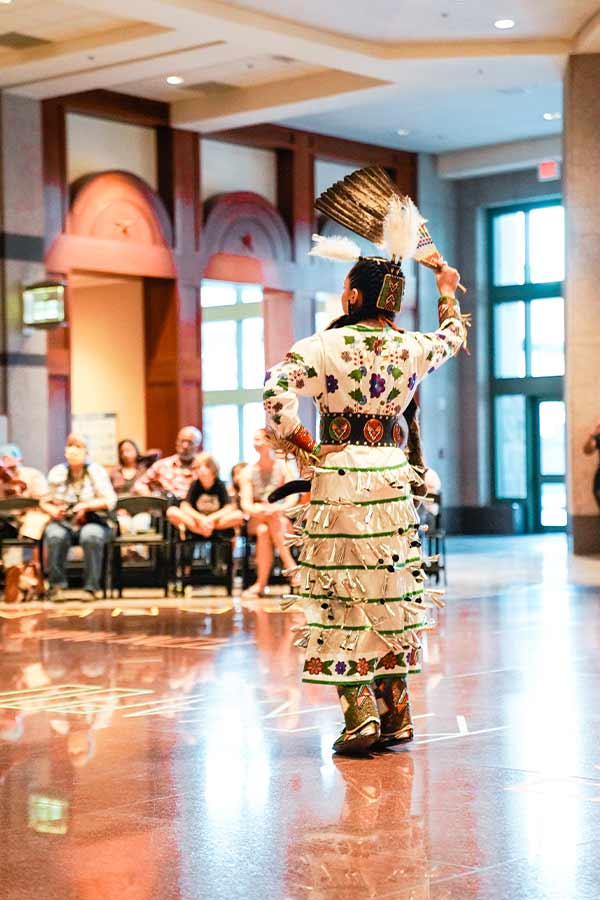 a Native American woman wearing a white jingle dress and holding a feather during a dance held in the Bullock Museum Grand Lobby