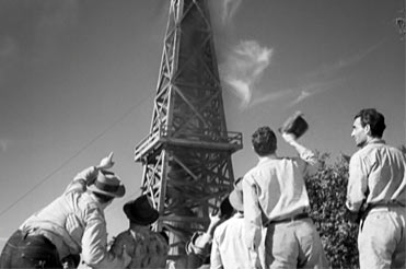 Black and white photo of a group of men looking up at an oil rig