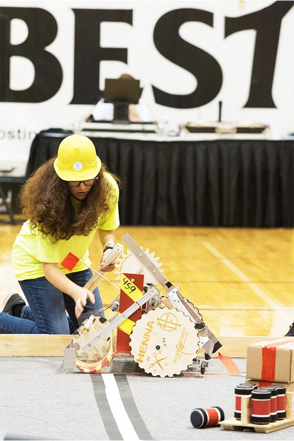 student with long hair and a hard hat crouched near a DYI robot at the Capitol BEST competition