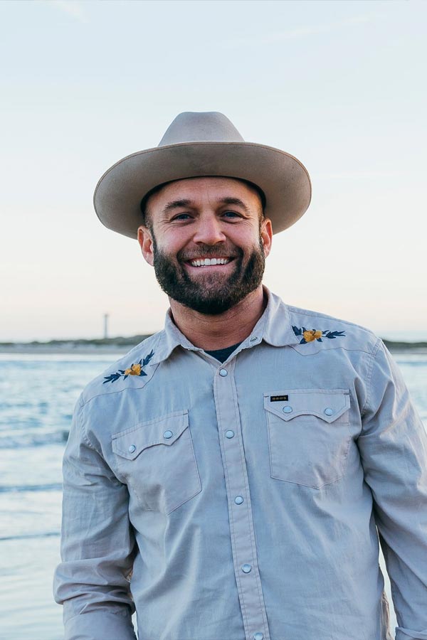 Chet Garner wearing a cowboy hat and denim shirt in front of a beach scene