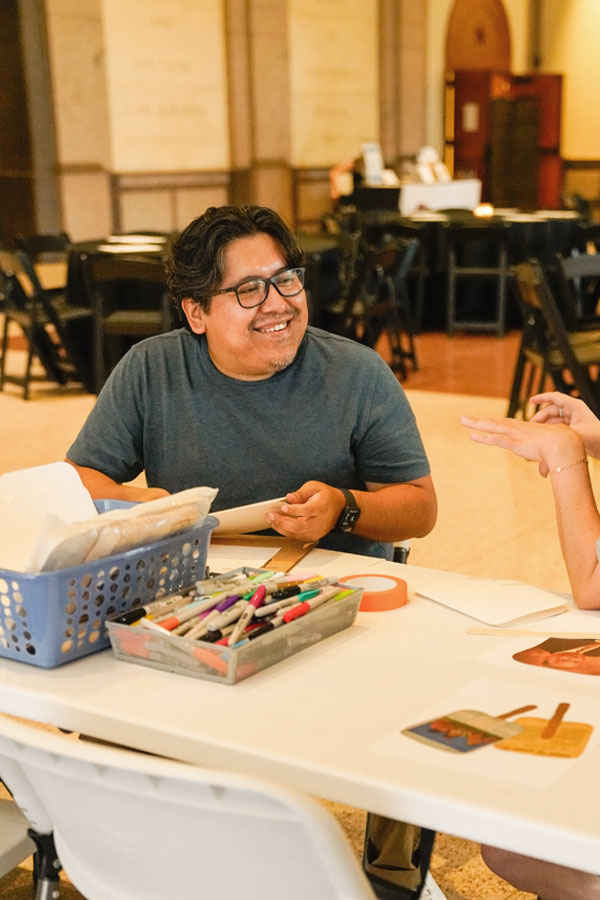 An adult sitting at a craft table and conversing with another adult off-camera.