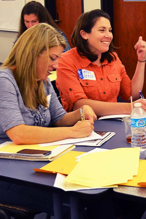 two women sitting at a table with worksheets in front of them, one woman is smiling, the other is looking down at her paper