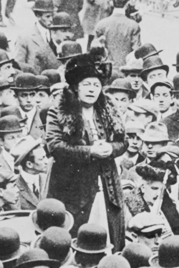 A black and white photo of a lone woman standing above a group of men, addressing the crowd in a street. All are wearing early 1900s attire.