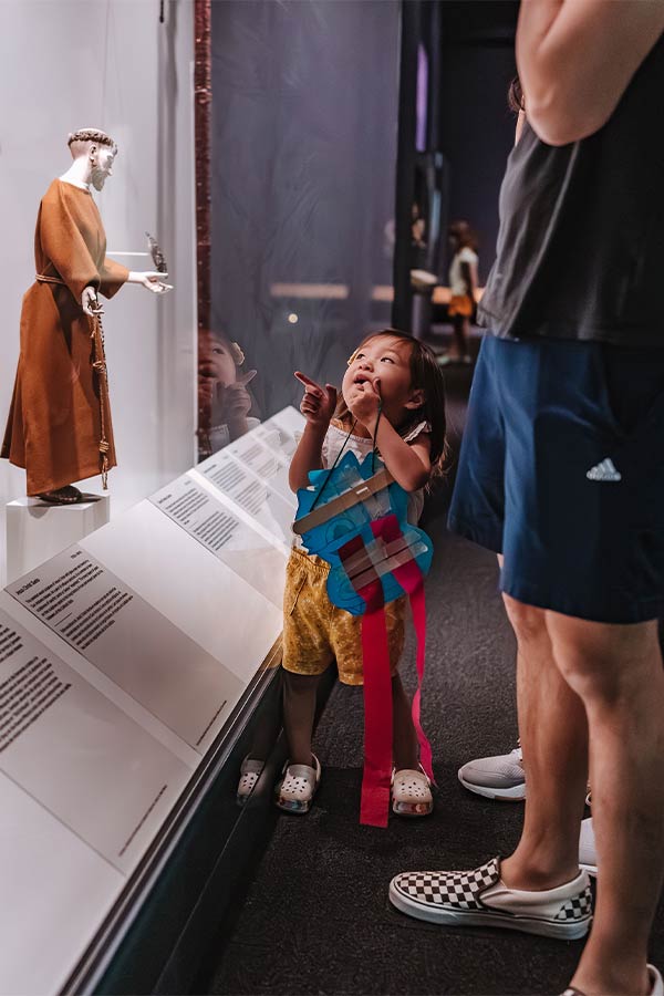 a young girl holding a craft while pointing up at a statue in a display case in the Bullock Museum