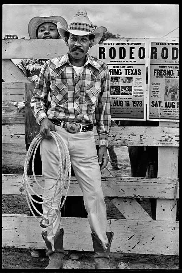 black and white photo of a man in a cowboy hat holding a lasso standing in front of a Texas Rodeo poster 
