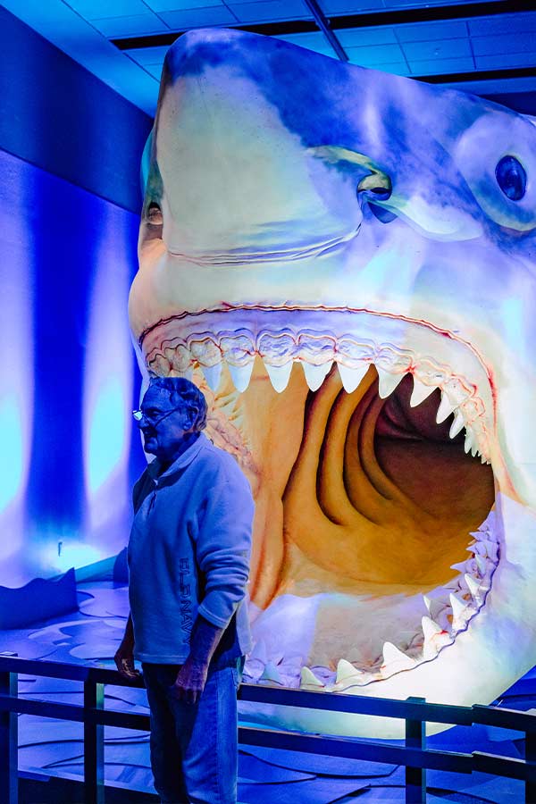 a man standing in front of a large model of a Megaladon shark with its mouth open