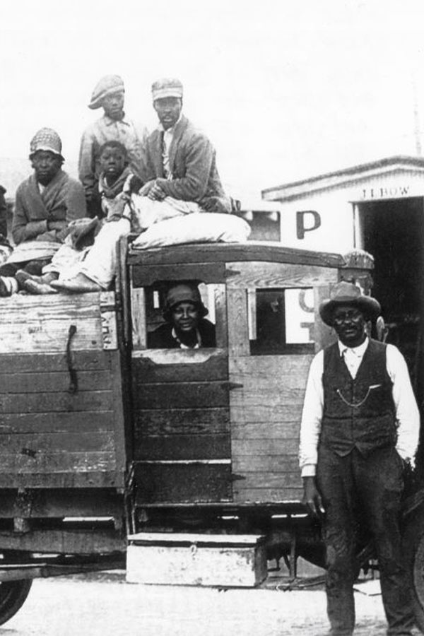 black and white photo of group of people sitting in the bed of an old truck with a man standing in front of it