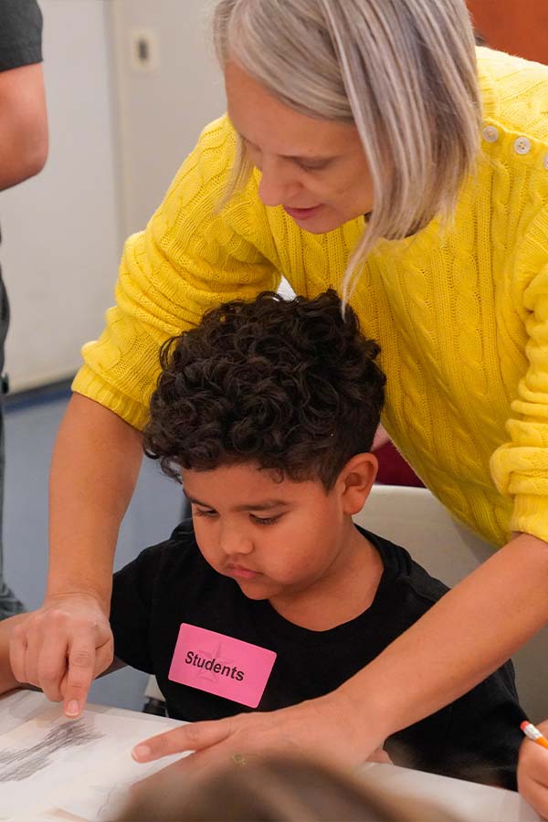 a young boy doing an drawing activity with a women in a bright yellow sweater behind him