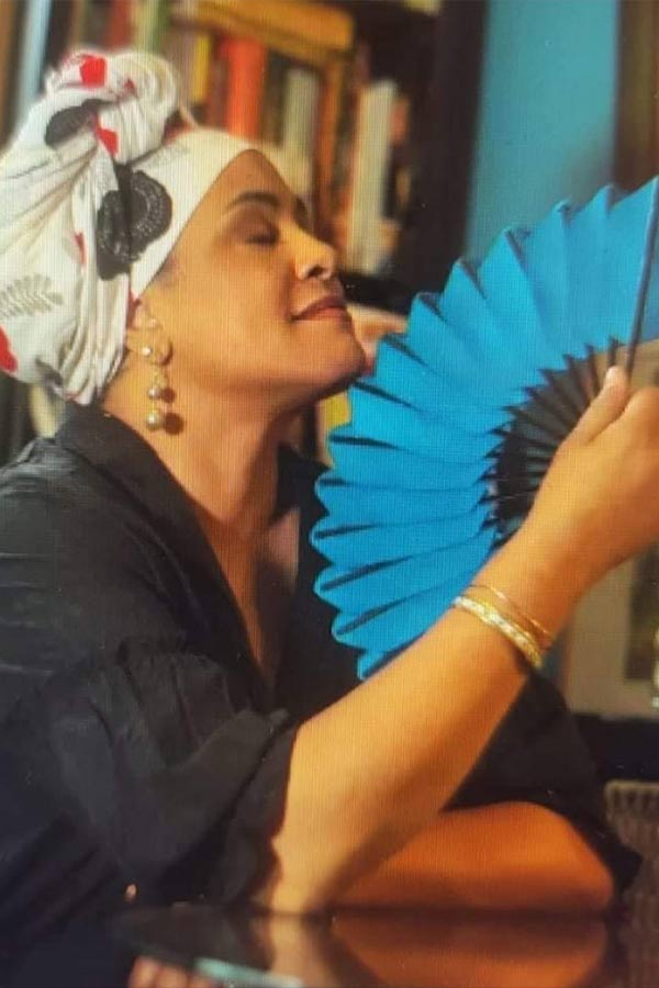 Halima Taha holding a blue fan, sitting in front of a bookcase