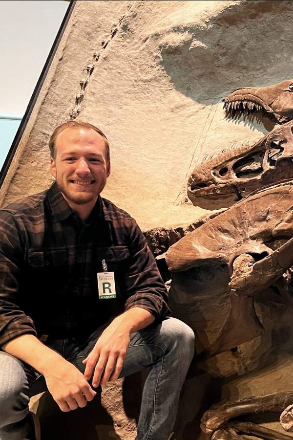 A man posing next to a wall of dinosaur fossils.