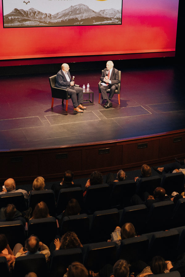 Two people sitting in chairs on a stage in a full theater.