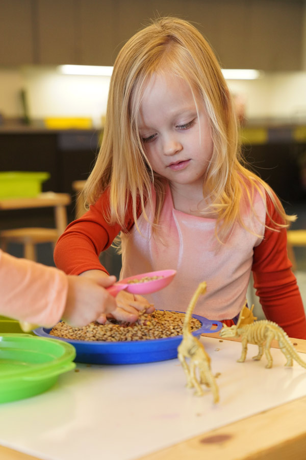 A child sitting at a craft table doing a digging activity with dinosaur figurines.
