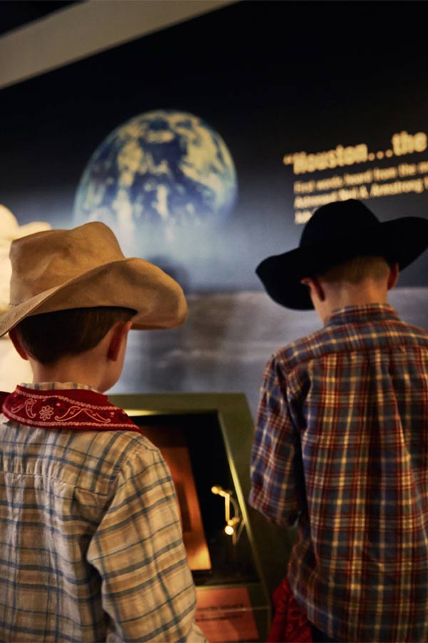 two children in plaid shirts and cowboy hats standing in front of a graphic of the Earth in the Bullock Museum