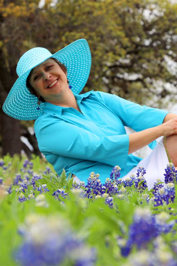 Woman posing as Ladybird Johnson wearing blue outfit and blue sun hat, sitting in a field of blue bonnet flowers