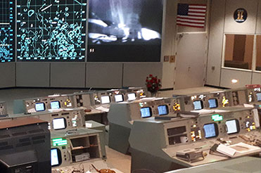 Interior of Mission Control Houston desks with computer consoles are the forefront and illuminated maps on the wall