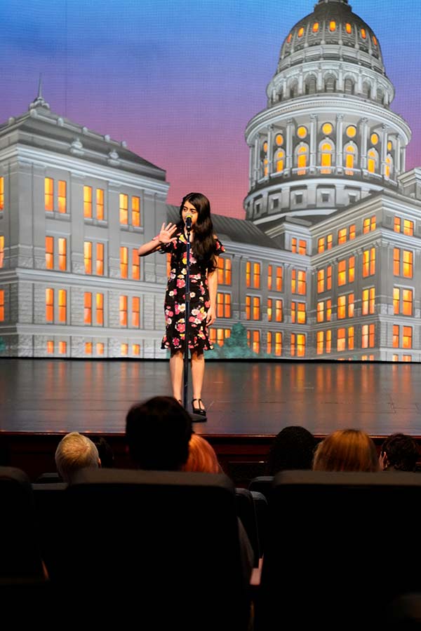 a young girl on a stage in front of a microphone with the Texas Capitol in the background