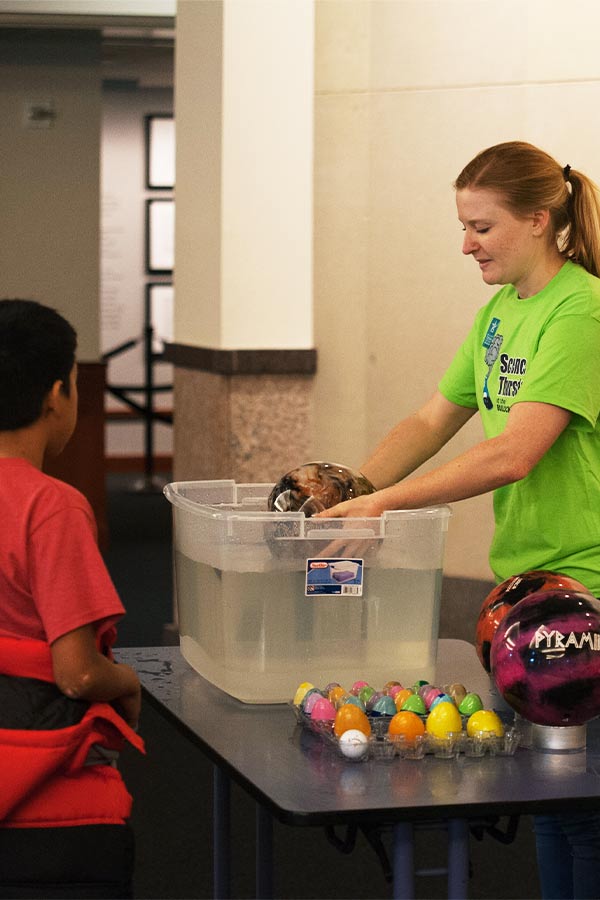 a woman in a green shirt and students gathered around a table. The woman is holding a bowling ball in a container of water.