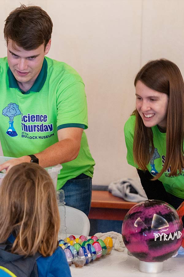 two adults wearing bright green shirts talking to a student at a table at Science Thursday 