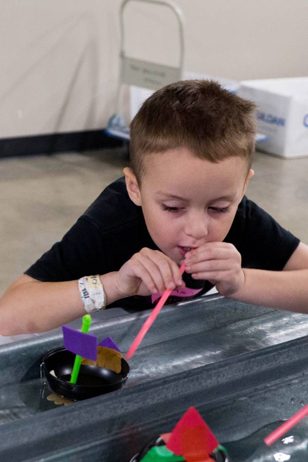 young boy blowing through a straw to make a paper boat move