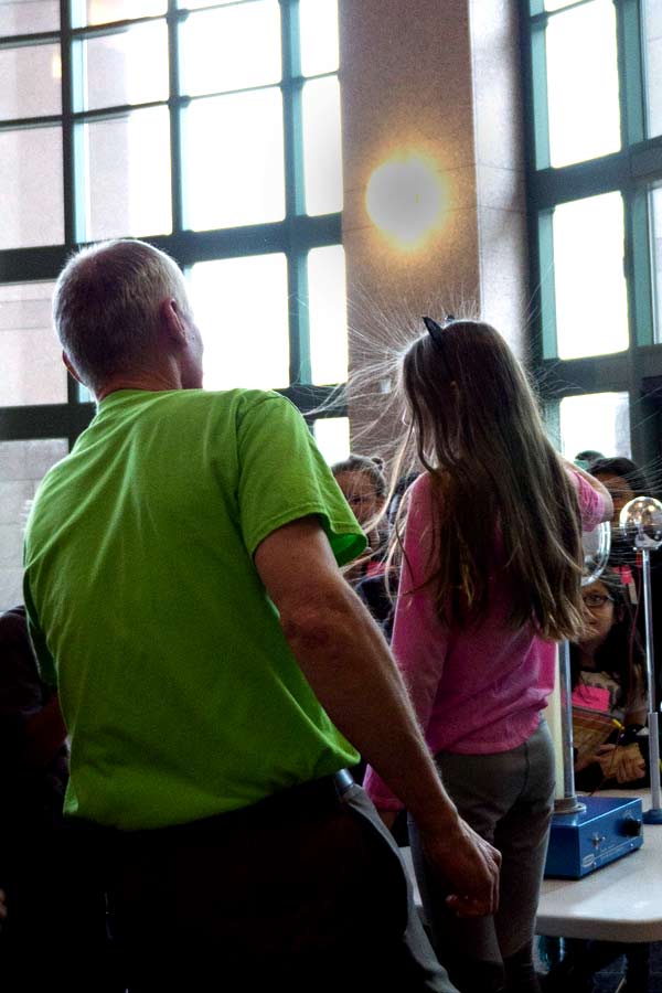 a man in a green shirt and a young student touching a Van Der Graff generator, the student's hair is standing up