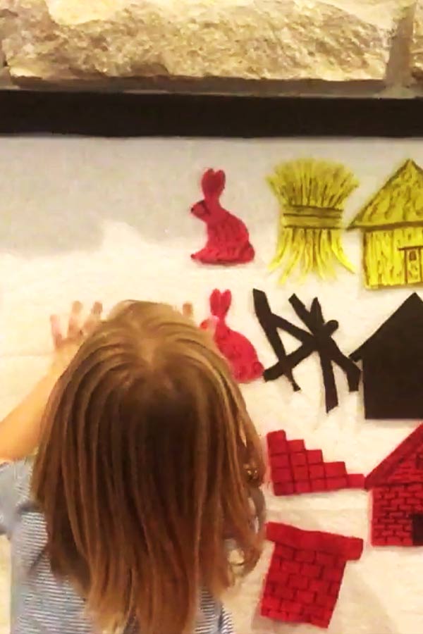 Young child placing a red rabbit on a white piece of paper on a wall. There are cut outs of bricks, a house, hay, and sticks also on the paper. 