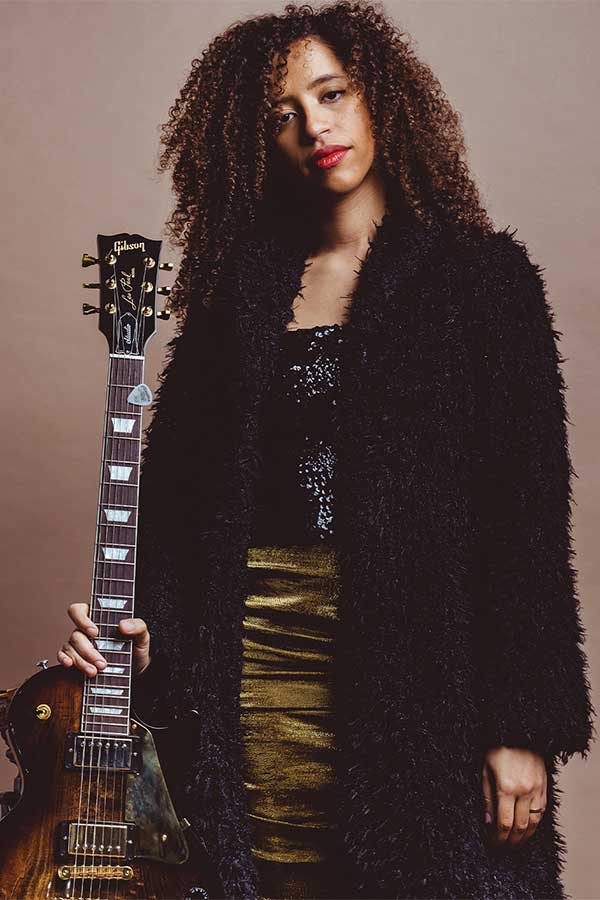 a woman standing with a stoic expression, propping up a guitar with her left hand