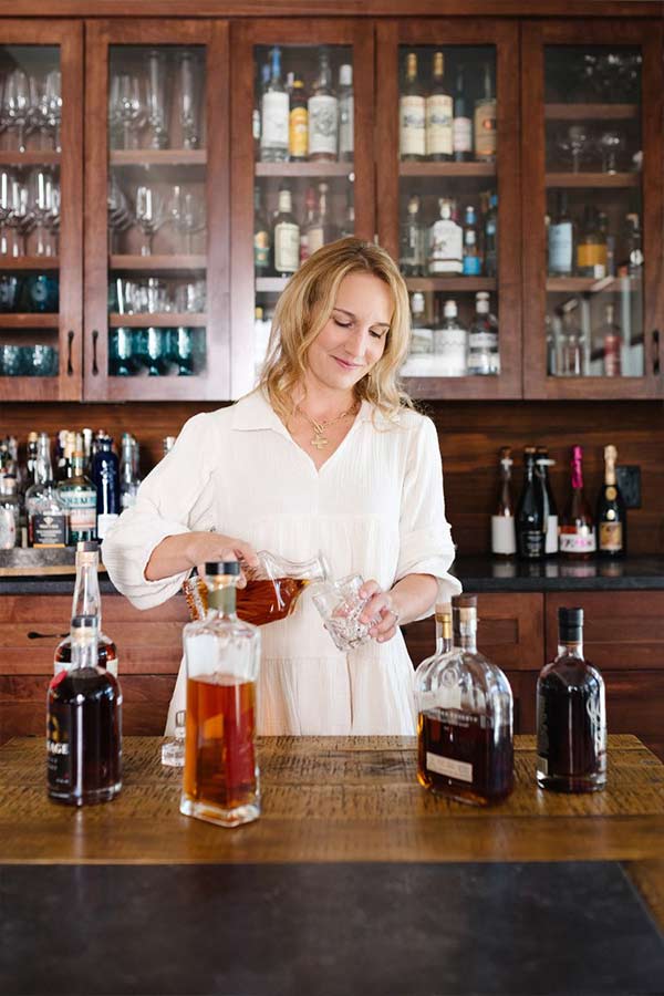 a woman standing behind a bar, pouring a drink with many different kinds of liquor on the counter
