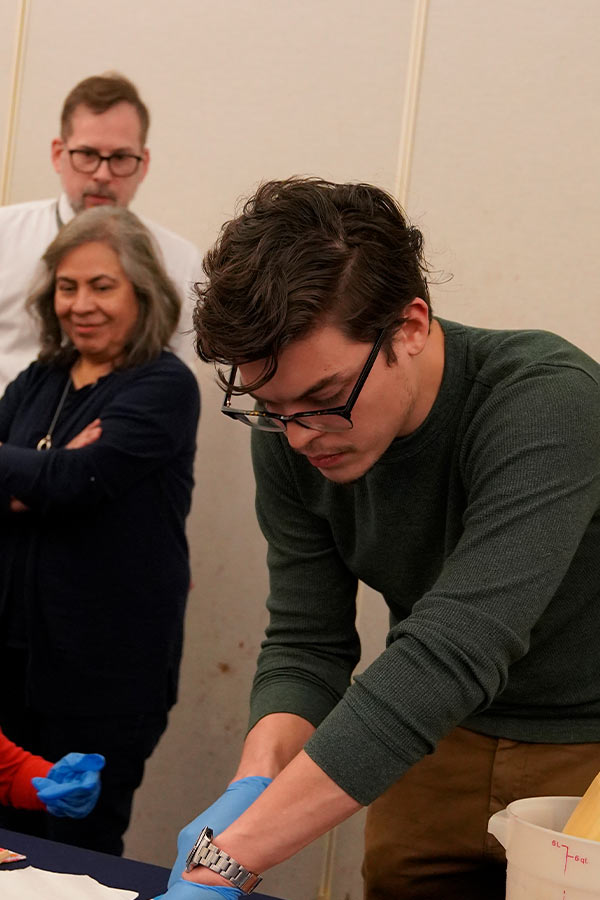 a young man with thick glasses bending over a table making something with his hands