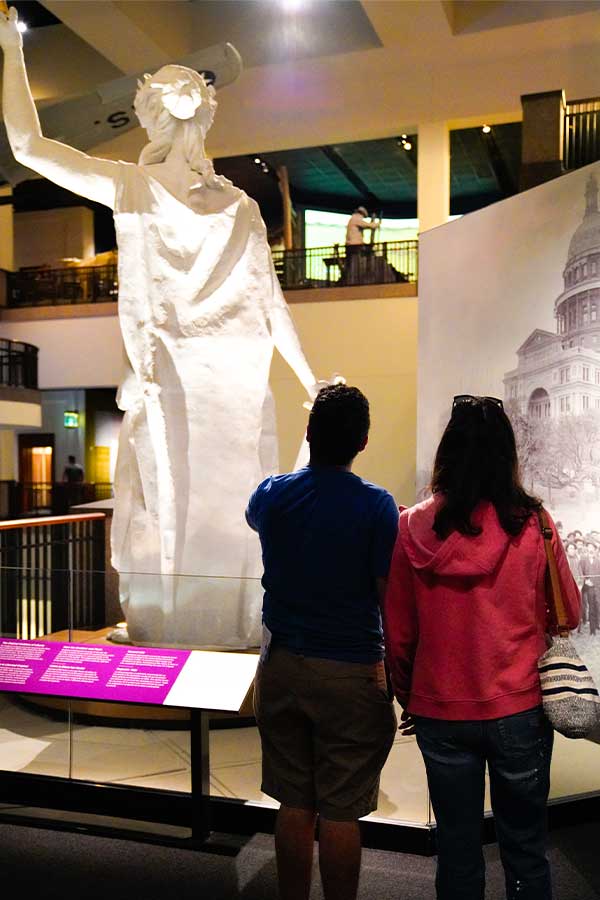 two adults looking at a white statue of the Goddess of Liberty