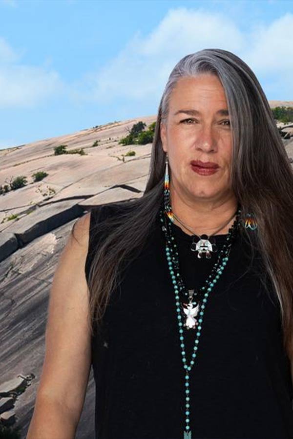 a woman with long gray hair standing in front of a large rock