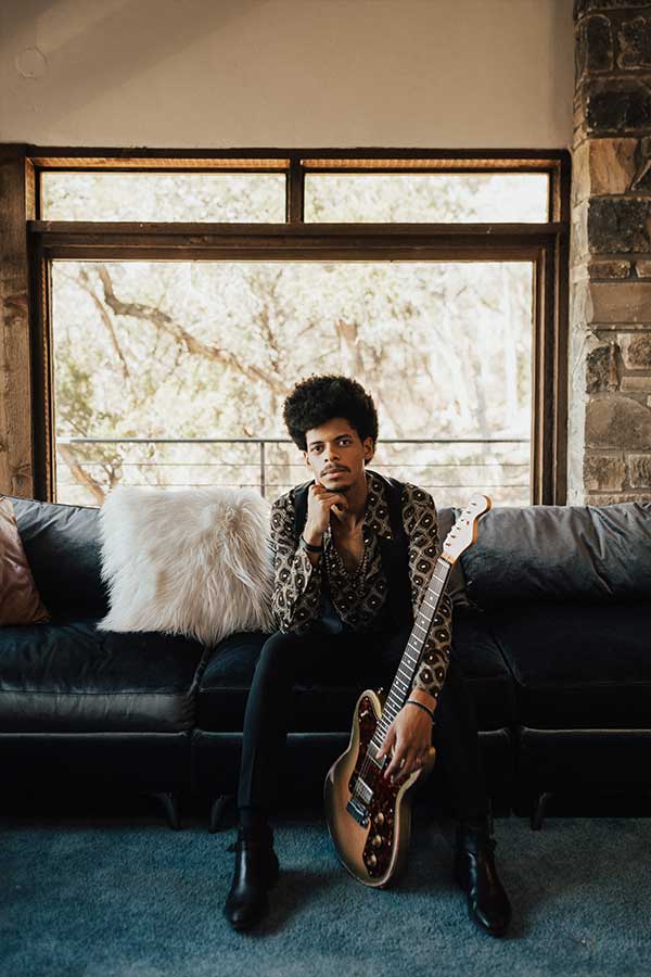 a man looking into the camera, sitting on a black couch, holding an electric guitar 