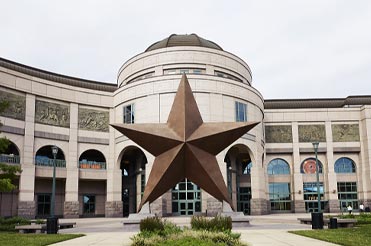 Exterior of Bullock Museum made of brown and red granite with a green dome and a large bronze star in the plaza