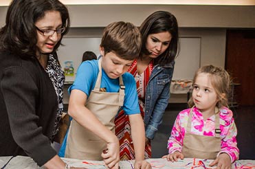 two children wearing brown aprons, using a stamp with two adults at the Bullock Museum