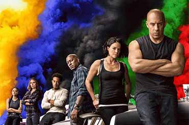 cast of F9 The Fast Sage standing in a row with Vin Diesil in the forefront, behind them is a colorful cloud of smoke