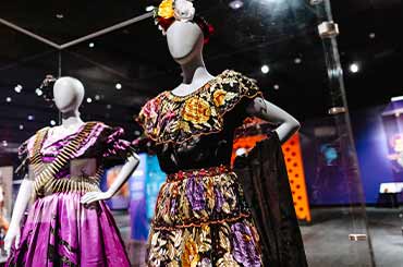 two traditional Mexican dresses on display in the Bullock Museum, the left is purple with a bullet studded sash and the right is a colorful floral pattern