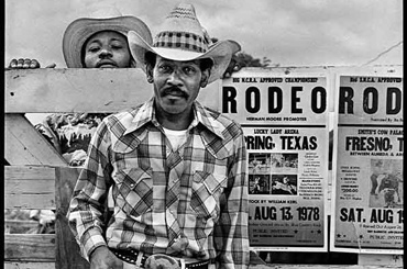 black and white photo of a man in a cowboy hat holding a lasso standing in front of a Texas Rodeo poster 