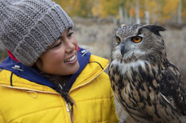 woman wearing a yellow jacket and grey hat with an owl perched on her arm