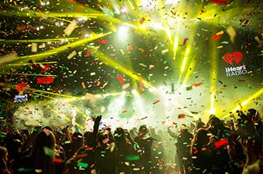 a stage with bright yellow lights beaming in all directions, confetti falls on a large audience