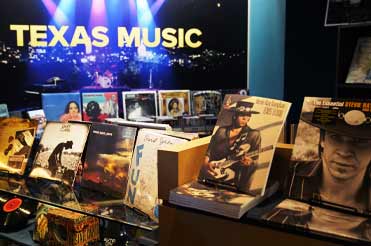 Assorted CDs and books on Texas music in the Bullock Museum Store, in the background is a lit up blue sign that reads TEXAS MUSIC