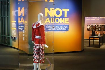 mannequin wearing a red Jingle Dress in a display case, in the background is a wall graphic that reads, "NOT ALONE"