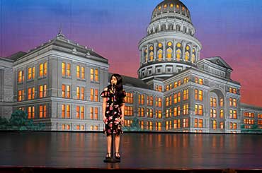 a young woman standing in front of a microphone on a stage with a projection of the Texas Capitol behind her