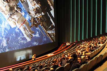people sitting in a large IMAX Theatre, an astronaut in space is on the screen