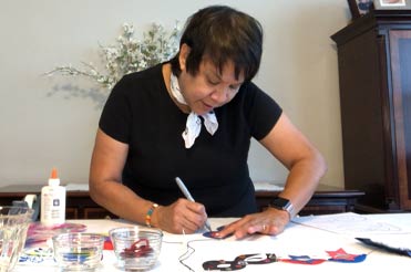 Woman standing at table with craft supplies, bent over drawing on paper