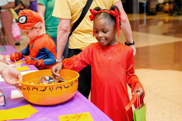 A child dressed in a costume gathering candy from a table.